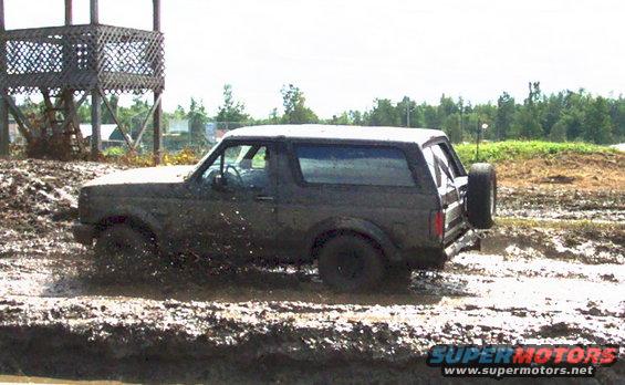 Muddy 1978 Ford Bronco driving through a mud pit with a wooden structure in the background