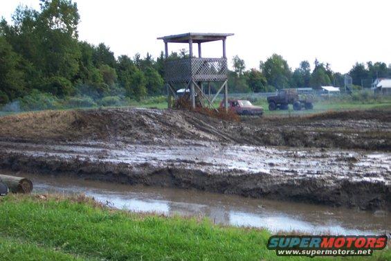 Mud bog with two trucks, one red and one dark, near a wooden observation tower. Muddy terrain and water are visible