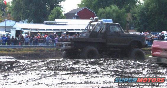 Modified pickup truck with large tires driving through mud, crowd watching in background