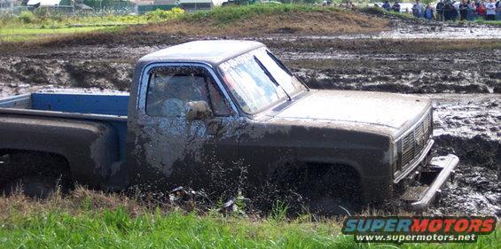 Mud-covered pickup truck stuck in a mud pit, 3/4 front view. Grille suggests 1980s model