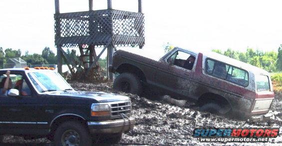 Ford Bronco stuck in mud next to a pickup truck, with a wooden structure in the background