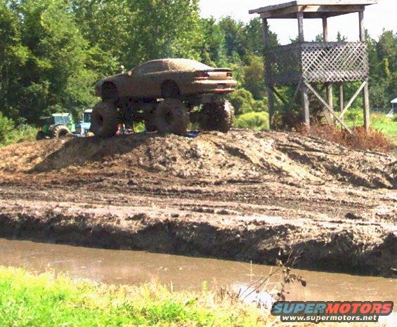 Mud-covered car on monster truck tires on a dirt mound next to a wooden observation tower. Tractor in background