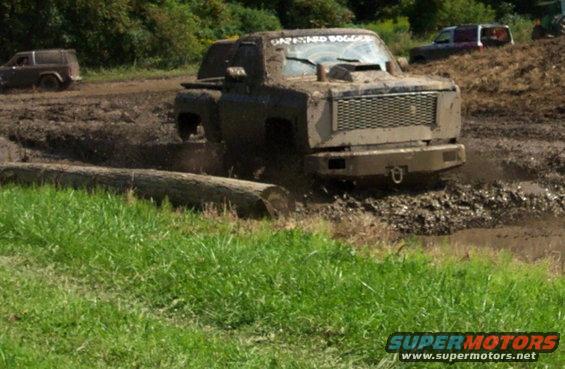 Mud-splattered pickup truck with a large grille and hood scoop stuck in mud, with a log in the foreground