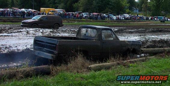 Mud bogging event with multiple trucks and SUVs stuck in mud, spectators in background