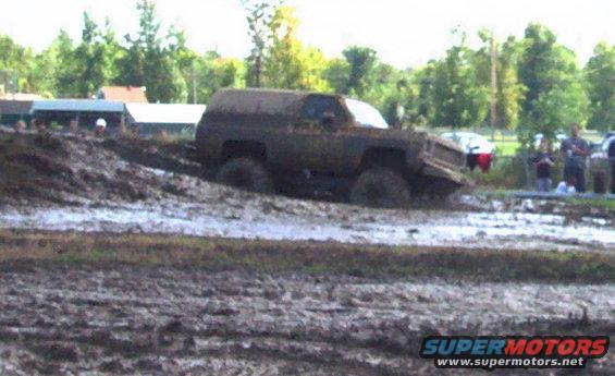 Muddy SUV stuck in a mud pit with trees and people in the background