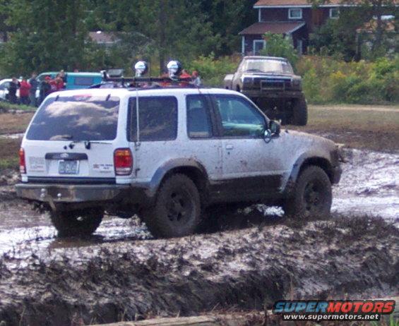 White SUV with roof rack and lights stuck in mud, another truck in background
