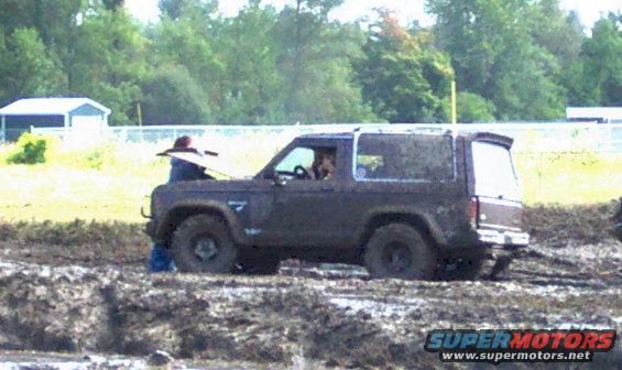 Dark brown Ford Bronco stuck in mud with hood open, person leaning over engine