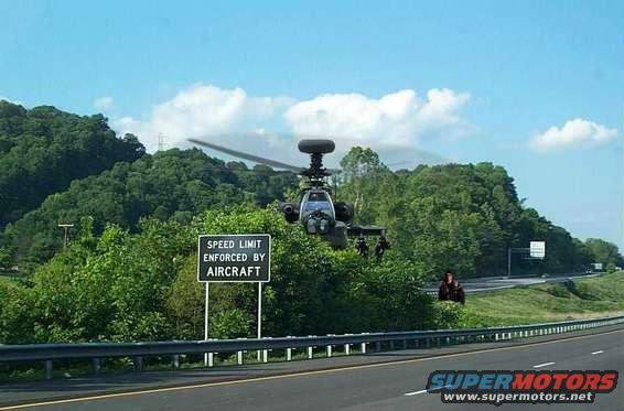 Apache helicopter flying low near a highway with a 'Speed Limit Enforced by Aircraft' sign