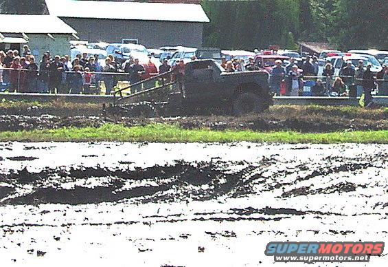 Dark pickup truck stuck in mud, side view. Spectators watch from a raised platform