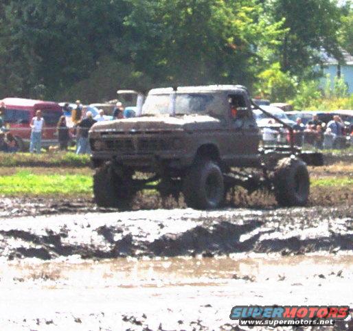 Mud-covered pickup truck with large tires driving through mud, front grille visible