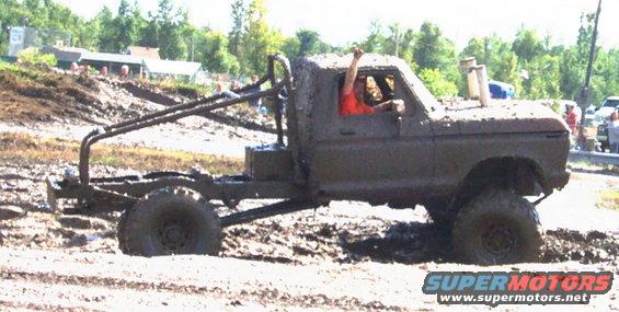 Mud-covered 1979 Ford Bronco with a roll cage and snorkel driving through a mud pit. Driver has arm raised