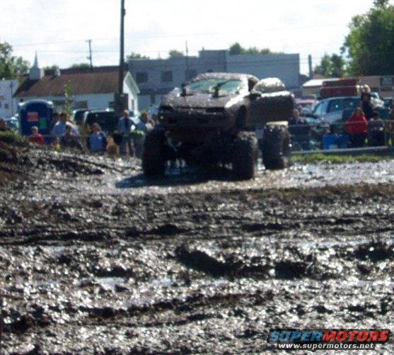 Monster truck with huge tires driving through mud, people watching in background