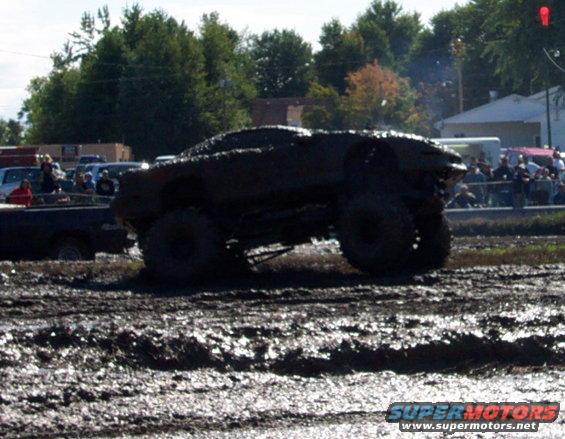 Mud-covered off-road vehicle with large tires driving through a muddy track, spectators in background