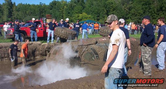 Mud-covered vehicle overturned in a mud pit with steam rising, onlookers present