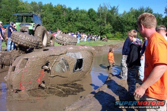 Mud bogging event with an overturned vehicle stuck in the mud, a tractor nearby, and spectators watching