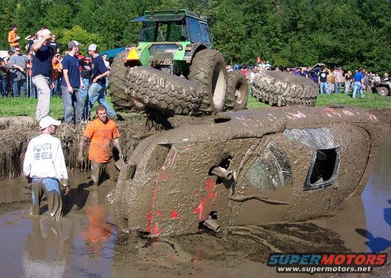 Muddy tractor on top of a submerged vehicle in a mud pit with spectators