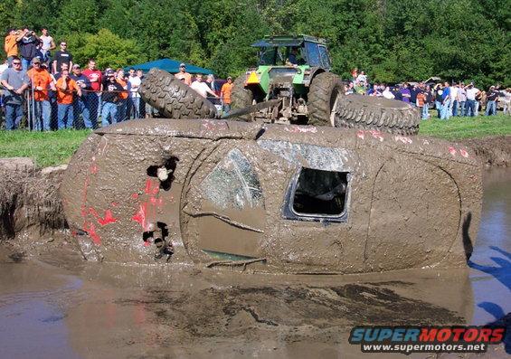 Mud-covered vehicle on its side, with a tractor behind it and spectators watching