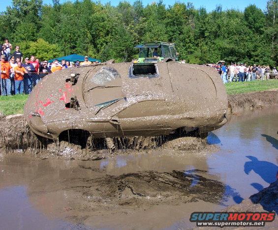 Mud bogging event with a heavily mud-covered vehicle stuck in mud, spectators watching, and a tractor in the background