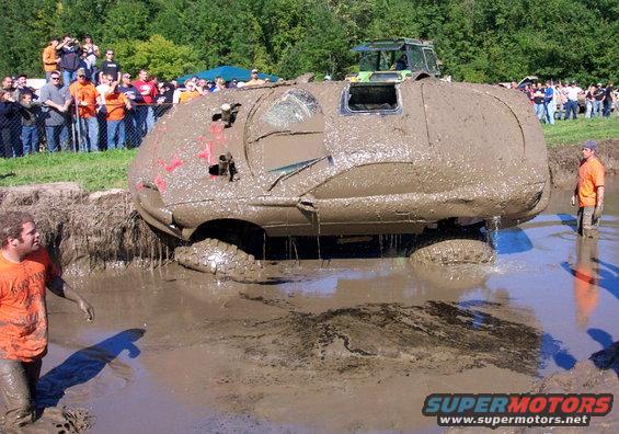 Mud-covered SUV stuck in a bog pit with spectators watching. Another vehicle is visible in the background