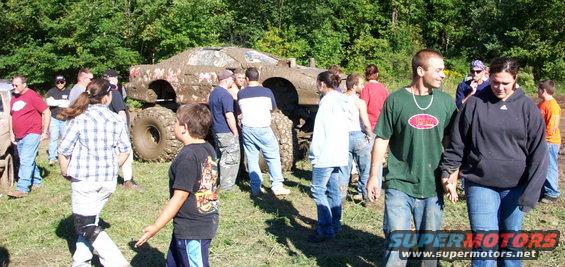 Muddy off-road vehicle with large tires, surrounded by people in a grassy, wooded area