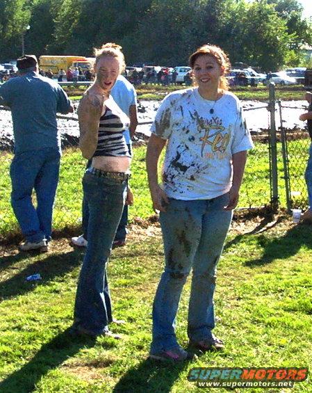 Two women covered in mud stand in a grassy field with vehicles and trees in the background
