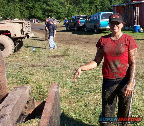 Muddy woman in red shirt and black pants, standing in a field with several vehicles in the background