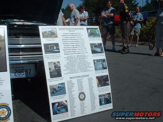 Display board listing sponsors and individuals at a car show, with a Washington license plate visible on a car in the foreground