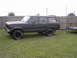 1977 Jeep Wagoneer - A dark-colored Jeep Grand Wagoneer is parked on a grassy lawn next to a wooden fence, attached to a small trailer.