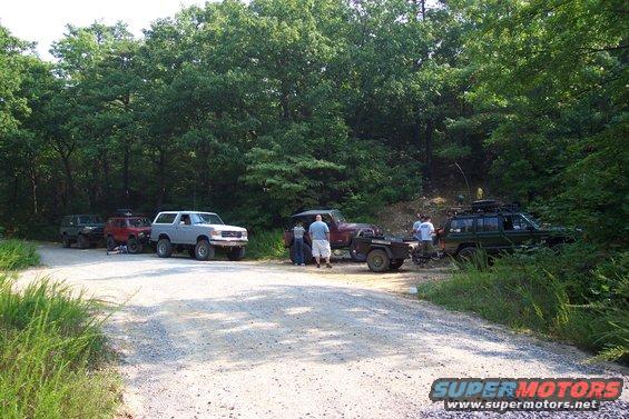 1987 Ford Bronco and other off-road vehicles parked on a dirt road with trees in the background