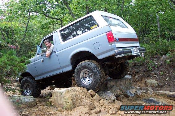 Light blue 1987 Ford Bronco with a white top, lifted suspension, and off-road tires, driving over rocks