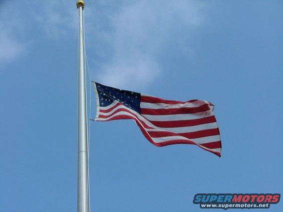 American flag flying on a flagpole against a blue sky. Supermotors.net logo in the corner