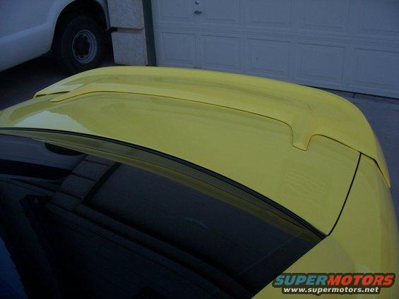 Yellow car spoiler and roof with a black rear window, a white truck is partially visible on the left