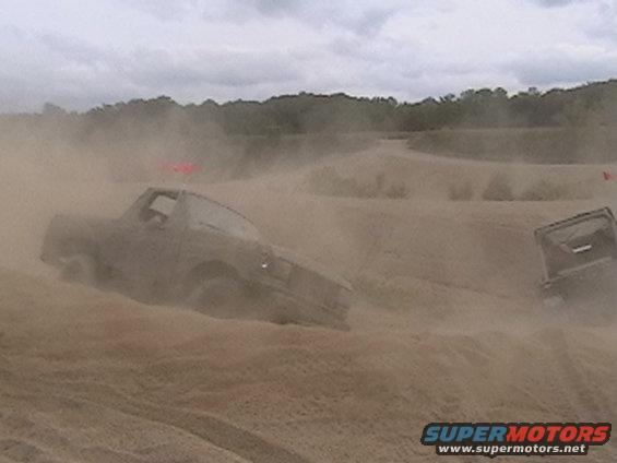 Truck kicking up dust on a dirt track, another vehicle visible in the background