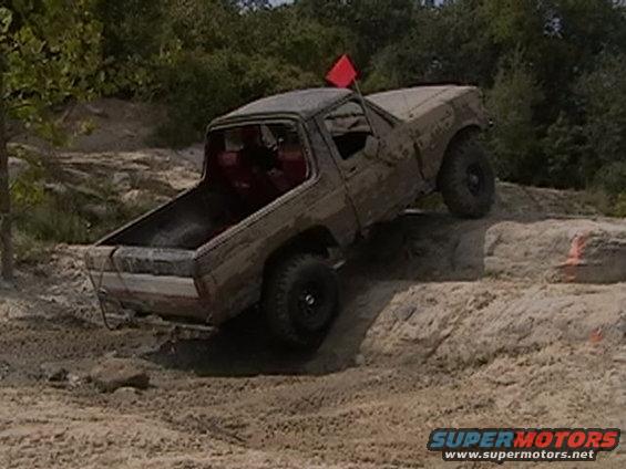 Brown pickup truck with red interior climbing a dirt mound, red flag on antenna. Appears to be a Ford F-series from the early 1980s