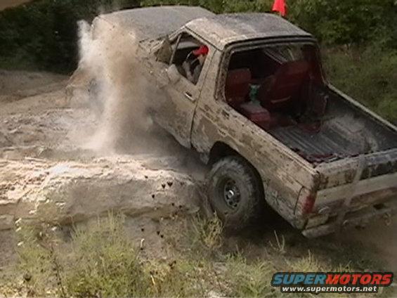 Mud-covered pickup truck navigating rocky terrain, kicking up dust. Red interior visible
