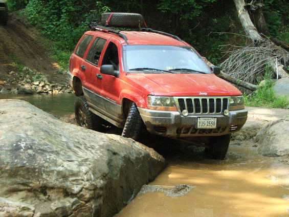 Red Jeep Grand Cherokee navigating muddy terrain with a spare tire on the roof rack