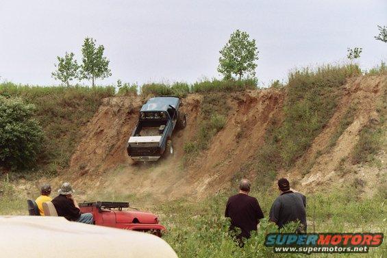 Blue pickup truck descending a dirt embankment, with spectators in a red vehicle below