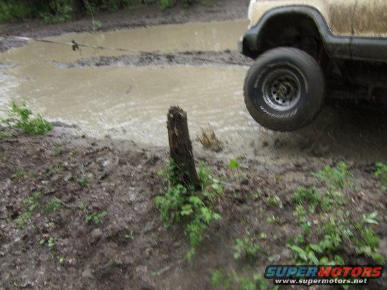 Off-road vehicle with one wheel lifted, driving through a muddy puddle. A wooden post is in the foreground