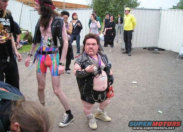 Two men in punk rock outfits, one with colorful shorts, stand outdoors with other people