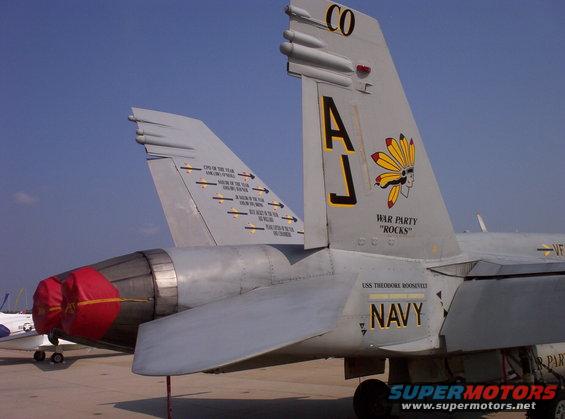 Two US Navy fighter jets parked on tarmac, one with 'AJ' and Native American headdress insignia, the other with mission tallies