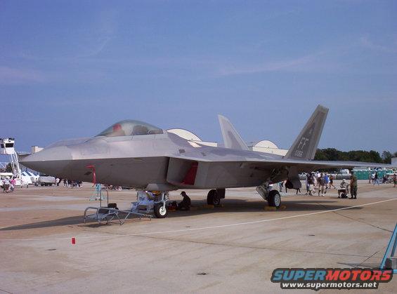 Gray fighter jet on tarmac with people around, under a clear blue sky