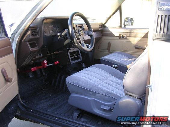 1985 Toyota 4Runner interior with grey cloth seats, aftermarket steering wheel, and a red steering wheel lock