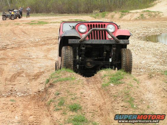 Red Jeep CJ-series driving over a dirt mound, front view. The vehicle has a prominent grille and round headlights