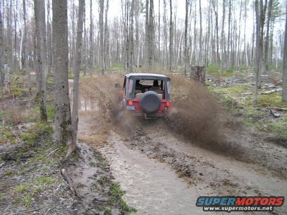 red Jeep Wrangler driving through mud and splashing water on a wooded trail