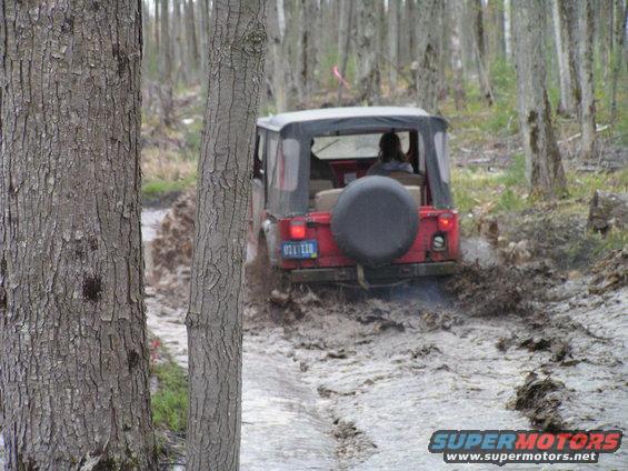 Red Jeep CJ-series vehicle with a spare tire on the back, driving through a muddy forest path