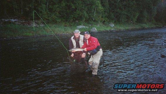Two men stand in a river, one holding a fish and a net, with trees in the background