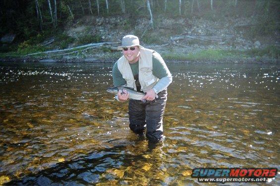 Guy in waders and a vest stands in a river holding a fish he caught. Trees and a rocky bank are in the background
