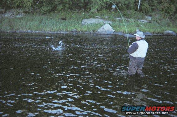 Man in waders casts fishing line into choppy river water with trees and rocks on the bank