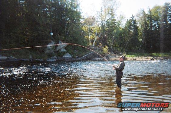 Man wearing waders and a hat, fly fishing in a river with trees and rocks in the background
