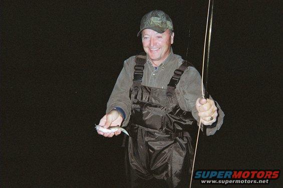 Man in fishing gear holds a small fish and a fishing rod against a dark background